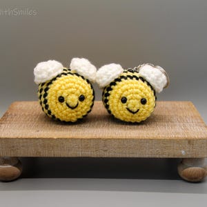 May include: Two crocheted bee keychains with smiling faces, yellow bodies with black stripes, and white wings. The keychains are displayed on a small, light brown wooden stand. The text "CrochetwithSmiles" is in the top left corner.
