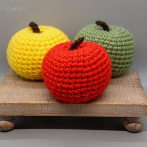 May include: Three crocheted apples on a wooden stand. The apples are red, yellow, and green.