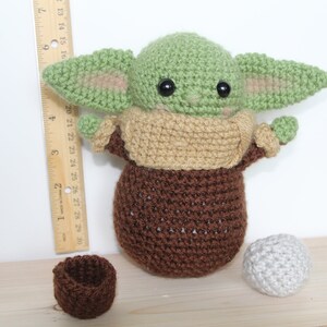 以下が含まれることがあります： スターウォーズのフランチャイズのキャラクターに似ている、緑と茶色の毛糸で編まれたぬいぐるみ。おもちゃは大きな目と小さな口をしています。おもちゃは、茶色の毛糸で編まれたボウルと灰色の毛糸で編まれたボールと一緒に、木製の表面の上に置かれています。