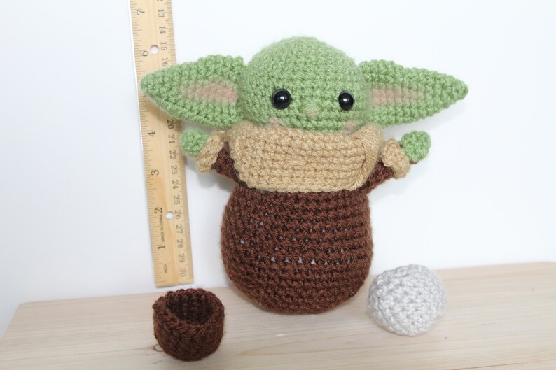 以下が含まれることがあります： スターウォーズのフランチャイズのキャラクターに似ている、緑と茶色の毛糸で編まれたぬいぐるみ。おもちゃは大きな目と小さな口をしています。おもちゃは、茶色の毛糸で編まれたボウルと灰色の毛糸で編まれたボールと一緒に、木製の表面の上に置かれています。