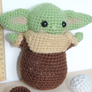 以下が含まれることがあります： スターウォーズのフランチャイズのキャラクターに似ている、緑と茶色の毛糸で編まれたぬいぐるみ。おもちゃは大きな目と小さな口をしています。おもちゃは木製の表面に置かれ、2つの小さな毛糸のボールがあります。