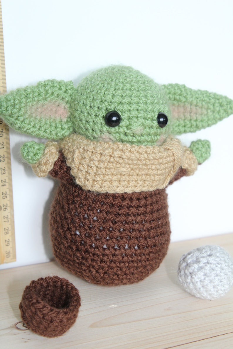 以下が含まれることがあります： スターウォーズのフランチャイズのキャラクターに似ている、緑と茶色の毛糸で編まれたぬいぐるみ。おもちゃは大きな目と小さな口をしています。おもちゃは木製の表面に置かれ、2つの小さな毛糸のボールがあります。