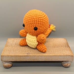 May include: Crocheted orange Charmander plush toy with black safety eyes and a yellow belly. The plush has a small flame tail with red and yellow accents. The toy is sitting on a small wooden display stand.