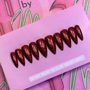 May include: A collection of red glitter press-on nails displayed on a pink card. The nails are stiletto-shaped and of varying sizes. The card features the text "by" and "USE CODE 'PRESSIO' FOR 10% OFF YOUR NEXT ORDER!"