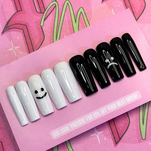 May include: A set of long, coffin-shaped press-on nails. Some nails are glossy black, others are white with a smiling face design. One nail features a sad face. The nails are displayed on a pink card with text.