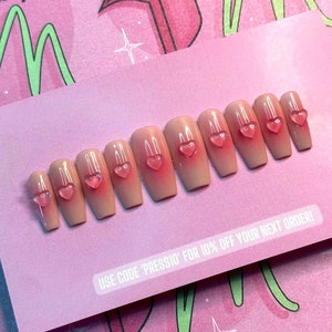 May include: A set of ten artificial nails in a light peach color with a gradient effect and small, clear pink heart-shaped embellishments. The nails are arranged on a pink card with text that reads "USE CODE 'PRESSIO' FOR 10% OFF YOUR NEXT ORDER!"