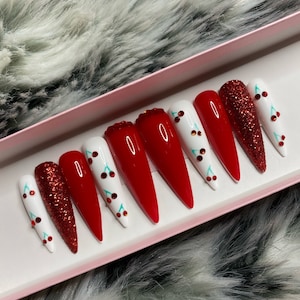 May include: A set of long, stiletto-shaped press-on nails. The nails feature a variety of designs, including solid red, red glitter, and white with cherry accents. The nails are displayed in a pink box against a fluffy, gray and white background.