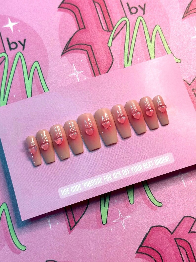 May include: A set of ten artificial nails in a light peach color with a gradient effect and small, clear pink heart-shaped embellishments. The nails are arranged on a pink card with text that reads "USE CODE 'PRESSIO' FOR 10% OFF YOUR NEXT ORDER!"
