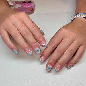May include: Manicured nails with a clear base, French tips, and a light blue accent. Some nails feature 3D flower designs and gold embellishments. The nails are on a light surface.