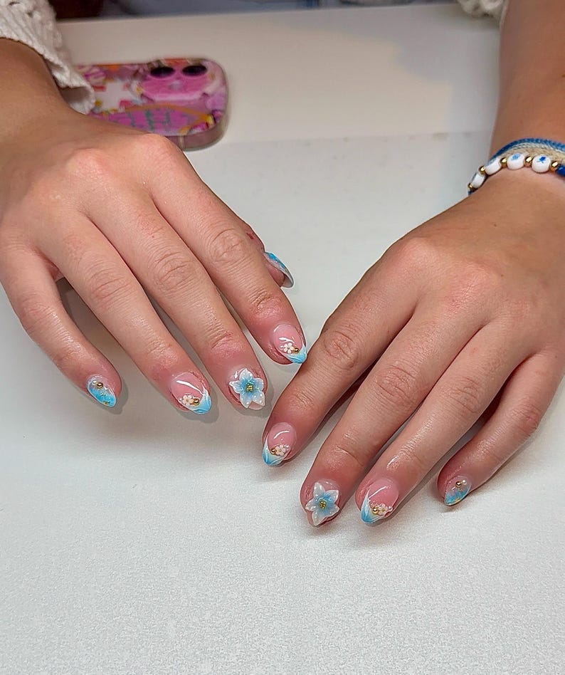 May include: Manicured nails with a clear base, French tips, and a light blue accent. Some nails feature 3D flower designs and gold embellishments. The nails are on a light surface.