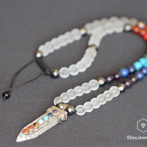May include: A clear quartz crystal pendant with seven chakra stones, set on a white frosted bead necklace with black hematite beads. The necklace has a black cord with black beads for adjusting the length.