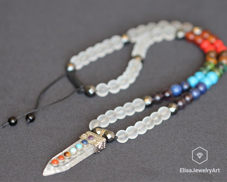 May include: A clear quartz crystal pendant with seven chakra stones, set on a white frosted bead necklace with black hematite beads. The necklace has a black cord with black beads for adjusting the length.