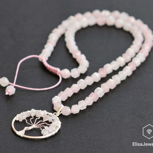 May include: A pink rose quartz necklace with a silver tree of life pendant. The necklace is made of faceted beads and has a pink cord with a heart-shaped charm.