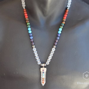 May include: A white crystal necklace with a silver pendant featuring seven chakra stones. The stones are red, orange, yellow, green, blue, indigo, and violet.