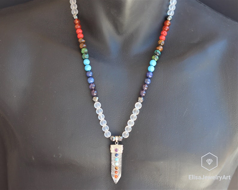 May include: A white crystal necklace with a silver pendant featuring seven chakra stones. The stones are red, orange, yellow, green, blue, indigo, and violet.