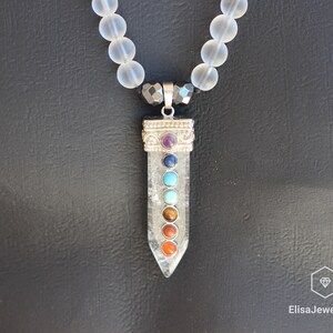 May include: A clear quartz crystal pendant with seven chakra stones, including amethyst, lapis lazuli, turquoise, green aventurine, tiger's eye, carnelian, and red jasper. The pendant is attached to a white frosted bead necklace.