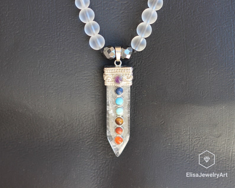 May include: A clear quartz crystal pendant with seven chakra stones, including amethyst, lapis lazuli, turquoise, green aventurine, tiger's eye, carnelian, and red jasper. The pendant is attached to a white frosted bead necklace.