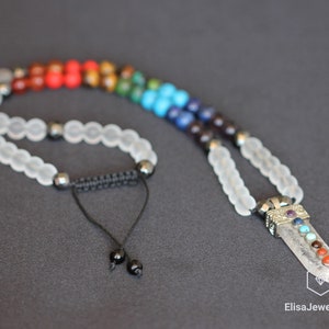 May include: A white crystal necklace with a chakra pendant. The pendant features seven gemstones in the colors of the chakras, red, orange, yellow, green, blue, indigo, and violet. The necklace is made with white beads and a black cord.