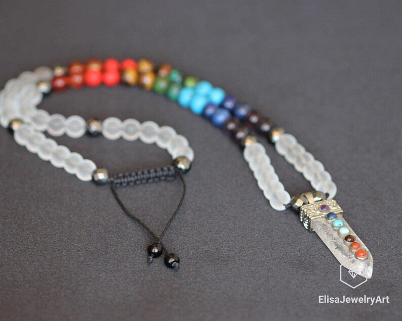 May include: A white crystal necklace with a chakra pendant. The pendant features seven gemstones in the colors of the chakras, red, orange, yellow, green, blue, indigo, and violet. The necklace is made with white beads and a black cord.