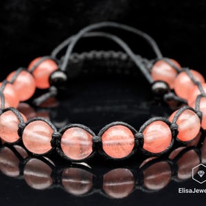 May include: A pink gemstone bracelet with black cord and knots. The bracelet is made with round, smooth gemstones.