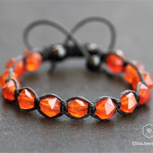 Puede incluir: Un brazalete de cordón negro con cuentas de piedras preciosas de color naranja. Las cuentas son facetadas y tienen una forma geométrica.