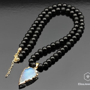 Puede incluir: Un collar de cuentas de ónix negro con un colgante de punta de flecha dorado con una piedra preciosa azul iridiscente. El collar tiene un cierre de mosquetón.