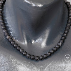 May include: A black beaded necklace with white marbling on each bead. The necklace is on a black mannequin.