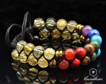 Puede incluir: Un brazalete de cuentas multicolores con un cordón negro. Las cuentas son de los colores del arcoíris, con separadores dorados entre cada color. El brazalete está atado con un nudo al final.