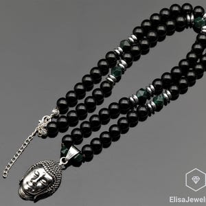 Puede incluir: Un collar de cuentas de ónix negro con un colgante de Buda de plata y detalles verdes. El collar está formado por dos hileras de cuentas.