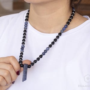 May include: A necklace made of black and blue beads with a long, rectangular pendant. The pendant is made of a blue stone.