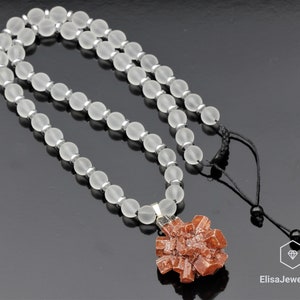 May include: A necklace made of frosted white beads with a pendant featuring a cluster of orange crystals. The necklace has a black cord with a silver clasp.