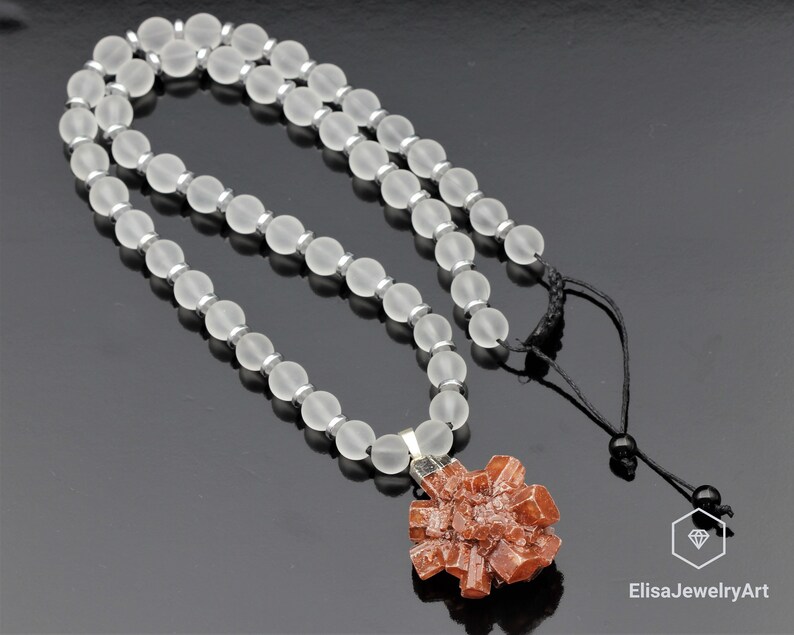 May include: A necklace made of frosted white beads with a pendant featuring a cluster of orange crystals. The necklace has a black cord with a silver clasp.