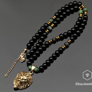 Puede incluir: Un collar de cuentas con esferas negras lisas, intercaladas con espaciadores metálicos dorados y acentos de piedra verde moteada. Un colgante de cabeza de león dorado cuelga del centro, unido a una cadena ajustable de tono dorado.