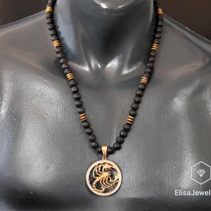 Op de afbeelding: Een zwarte lavastenen kralenketting met een gouden hanger in de vorm van een schorpioen. De hanger is omringd door heldere strass steentjes.