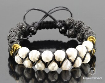 May include: A handmade bracelet featuring black lava rock beads, white gemstone beads, and gold-colored metal accents. The bracelet is strung on black cord and has an adjustable closure. The jewelry is displayed on a reflective surface.