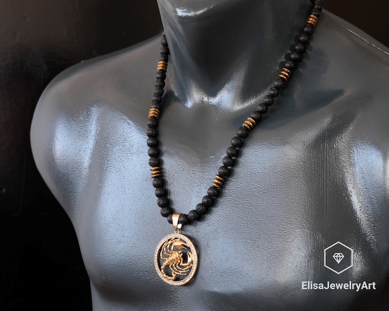 Op de afbeelding: Een zwarte kralenketting met een gouden hanger met een schorpioenontwerp. De ketting is om de nek van een mannequin.