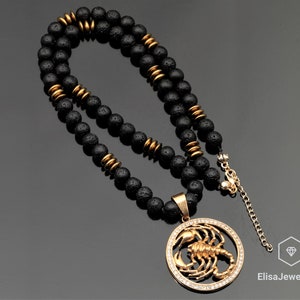 Op de afbeelding: Een zwarte lavastenen kralenketting met een gouden hanger met een schorpioenontwerp met heldere kristallen. De hanger is met een gouden ketting aan de ketting bevestigd.