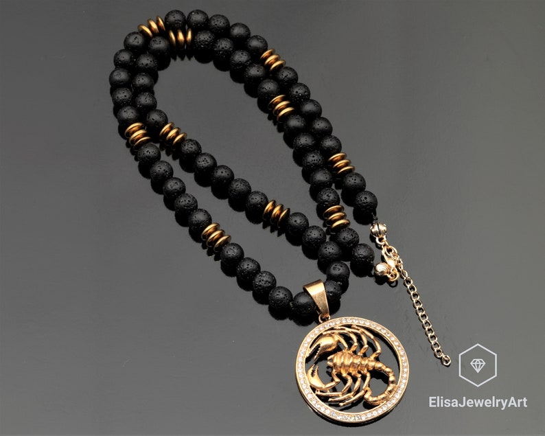 Op de afbeelding: Een zwarte lavastenen kralenketting met een gouden hanger met een schorpioenontwerp met heldere kristallen. De hanger is met een gouden ketting aan de ketting bevestigd.