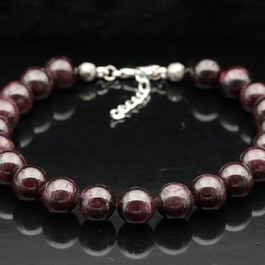 Puede incluir: Un brazalete de piedras preciosas rojas con un cierre plateado. El brazalete está hecho de cuentas redondas y tiene una longitud aproximada de 18 cm.