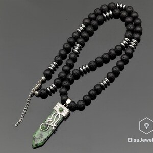 May include: A necklace featuring black porous lava beads interspersed with metallic silver-toned spacers. The pendant is a long, pointed green and black stone encased in a silver-toned metal cap with a wire-wrapped floral design and a small green bead.
