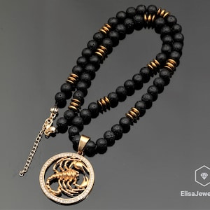 Op de afbeelding: Een zwarte lavastenen kralenketting met een gouden schorpioen hanger. De hanger is omringd door heldere strass stenen.