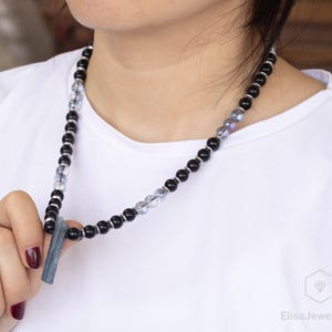 May include: A beaded necklace featuring black and iridescent beads, with a rectangular blue-gray pendant. The necklace is held by a person wearing a white shirt. The necklace is a piece of jewelry.