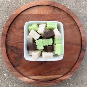 May include: A square white bowl filled with assorted wax melts in the shape of small blocks. The melts are in shades of white, light green, and brown. The bowl sits on a round, wooden serving tray.
