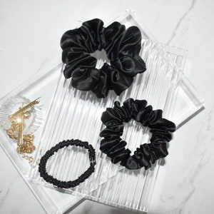May include: Four black satin scrunchies of varying sizes are arranged on a clear, textured tray. The scrunchies are made of a shiny, dark fabric. A small gold hair clip and chain are visible in the corner.