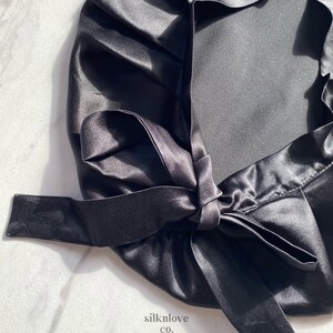 May include: A black satin bonnet with a tied bow closure. The bonnet is made of a shiny, smooth fabric and is designed to protect hair. The text "silknlove co." is visible in the lower right corner.