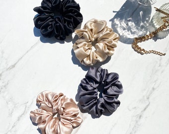 May include: Four scrunchies in different colors: black, beige, light pink, and dark gray. The scrunchies are made of a soft, silky material and are arranged on a white marble surface.