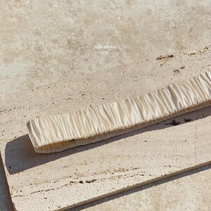 K&ouml;nnte beinhalten: Ein cremefarbenes Seidenstirnband mit gerafftem Design. Das Stirnband liegt auf einer strukturierten, hellfarbigen Oberfl&auml;che. Der Text "silknlove co." ist im Hintergrund sichtbar.