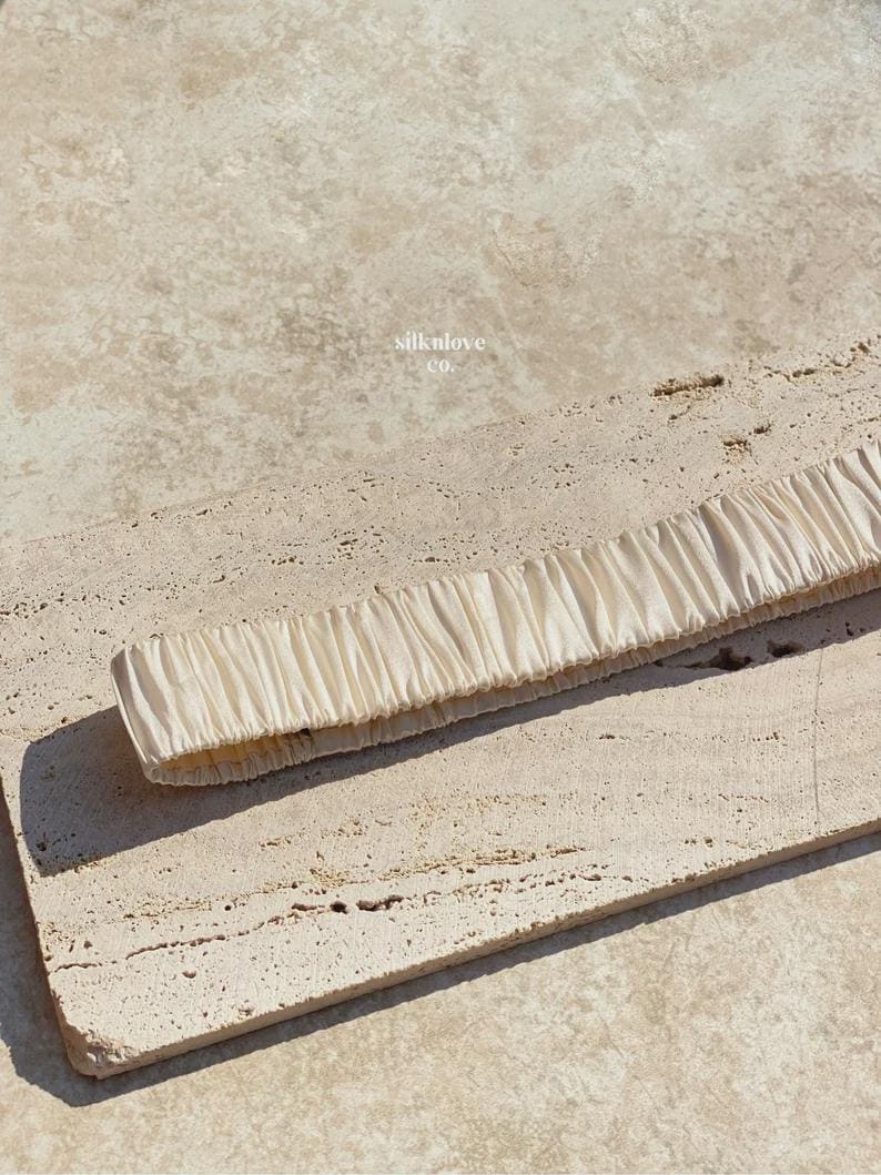 K&ouml;nnte beinhalten: Ein cremefarbenes Seidenstirnband mit gerafftem Design. Das Stirnband liegt auf einer strukturierten, hellfarbigen Oberfl&auml;che. Der Text "silknlove co." ist im Hintergrund sichtbar.