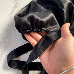 May include: A black satin bonnet with an elastic band and long ties. The bonnet is held in a hand, showcasing the smooth, shiny texture of the fabric. The brand name "silknlove co." is visible in the lower left corner.