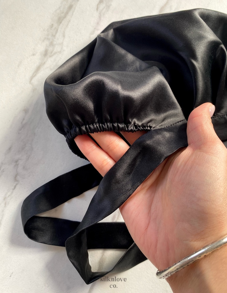 May include: A black satin bonnet with an elastic band and long ties. The bonnet is held in a hand, showcasing the smooth, shiny texture of the fabric. The brand name "silknlove co." is visible in the lower left corner.